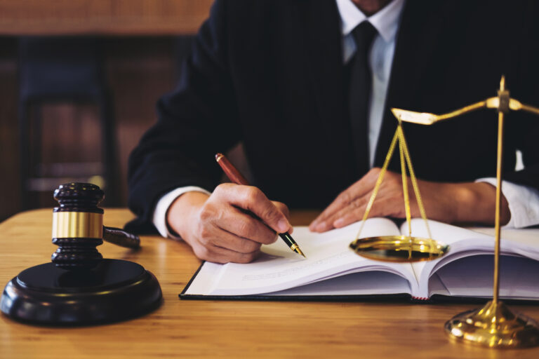 Image of a lawyer signing a document with the scales of justice and a gavel on his desk